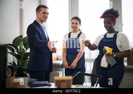 Un giovane uomo adulto caucasico che saluta due giovani donne adulte che puliscono, una caucasica e una nera, entrambe indossano uniformi e tengono in mano i prodotti per la pulizia, in piedi insieme in un ufficio moderno Foto Stock