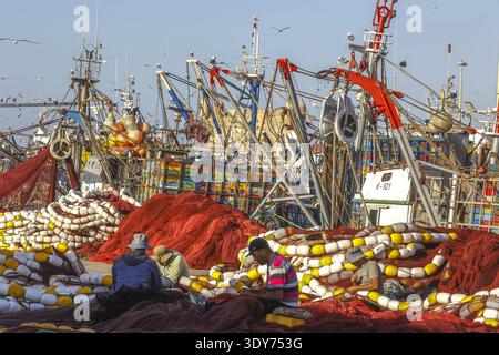 La mending di reti da pesca avviene nel porto con imbarcazioni e reti che circondano i lavoratori di Essaouira in Marocco Foto Stock