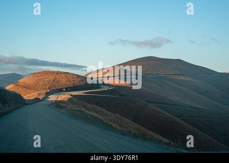 Solitaria strada di ghiaia che si snoda attraverso colline dorate sotto la luce del sole nel paesaggio neozelandese Foto Stock