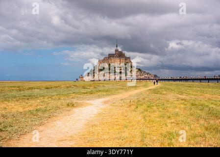 I visitatori camminano lungo il sentiero verso l'abbazia di Mont Saint Michel attraverso i prati della Normandia sotto il cielo nuvoloso Foto Stock