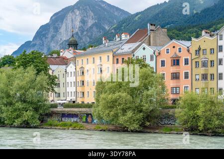 Edifici colorati si riflettono sul fiume Inn sotto un cielo soleggiato in estate con montagne sullo sfondo Foto Stock
