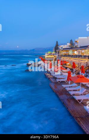 Taverna sul mare al crepuscolo con ombrelli arancioni a Limenas Chersonisou, Creta, Grecia Foto Stock