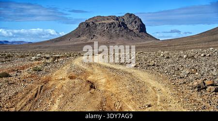 Strada sterrata che si snoda attraverso un vasto paesaggio desertico roccioso, che conduce verso un'imponente montagna dalla cima piatta sotto un cielo azzurro limpido con nuvole sparse, Foto Stock