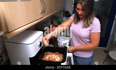 La donna prepara un piatto in una cucina moderna, aggiungendo formaggio a un vassoio di pasta all'interno di una friggitrice ad aria Foto Stock