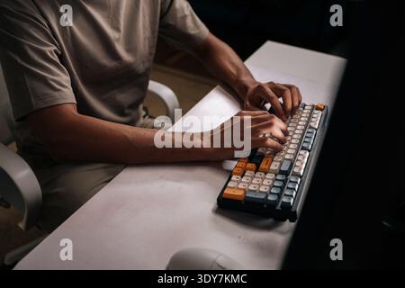 Foto ritagliata di tipizzazione maschile non riconoscibile da freelance su tastiera meccanica con tasti colorati in un ufficio buio, suggerendo un lavoro concentrato o tardi Foto Stock