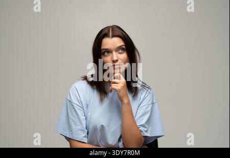 Giovane donna con t-shirt casual blu che guarda in alto, perde nel pensiero, contemplando un'idea o prendendo una decisione importante Foto Stock