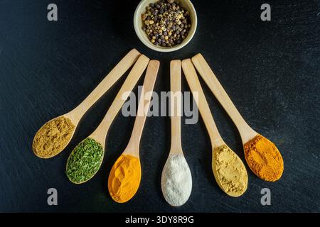 Spezie ed erbe assortite in cucchiai di legno su sfondo di ardesia scura con ciotola di grani di pepe, vista dall'alto, cucina e condimento. Foto Stock
