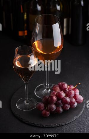 Due bicchieri di vino bianco e di rosa frizzante con grappoli di uva su una superficie scura di ardesia su bottiglie sullo sfondo Foto Stock