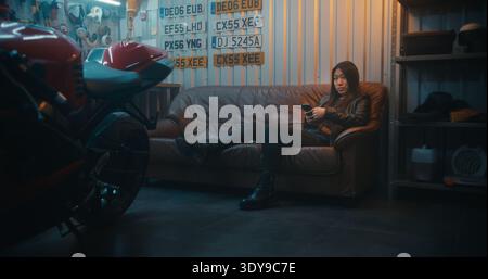 Donna sdraiata in garage con telefono e Sport Motorcycle. Donna in pelle nera sul divano marrone vede il video girato durante la corsa. Sottocultura per motociclisti e comunicazione moderna in garage privato. Verticale. Foto Stock