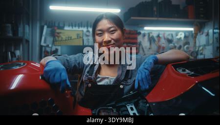 Ritratto di una donna felice sorridente che si appoggia alla motocicletta dopo una riparazione riuscita. Giovane donna asiatica soddisfazione dopo aver terminato la manutenzione tecnica e la riparazione elettronica in officina garage. Foto Stock