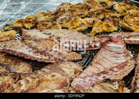 Una grande varietà di costolette di maiale condite e pezzi di pollo arrostiti su una griglia in metallo professionale all'aperto in un mercato alimentare. Foto Stock
