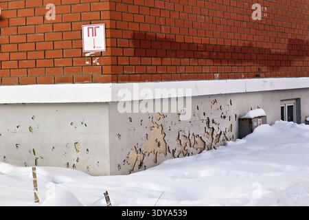 Zoccolo dell'edificio fatiscente con vernice sbucciante che rivela il calcestruzzo usurato. Parete in mattoni rossi sopra, a contrasto con neve bianca fresca che ricopre il terreno b Foto Stock