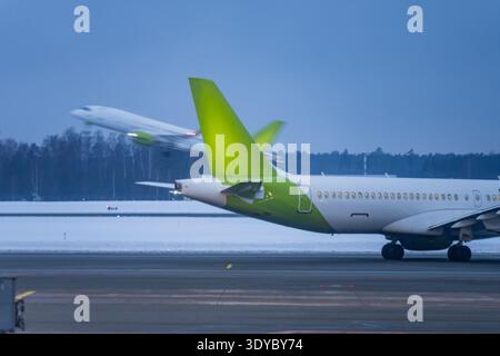 Jet commerciale con coda verde sul grembiule innevato dell'aeroporto durante l'ora blu, aereo sfocato in movimento in lontananza, illustrando gli orari dei voli e i traffici aerei Foto Stock