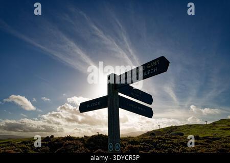 Il sole splende dietro un palo che mostra la direzione dei sentieri in un paesaggio collinare con un cielo blu e nuvole bianche, Great orme, Llandudno. Foto Stock