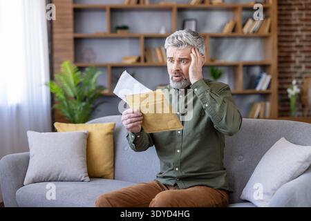 Uomo maturo seduto sul divano di casa, scioccato e stressato mentre apre una busta con cattive notizie finanziarie, bolletta inaspettata o avviso di debito, mano a testa, preoccupato e turbato Foto Stock