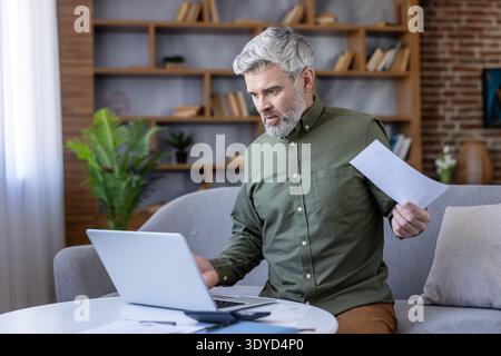 Uomo maturo con i capelli grigi che sembra scioccato e stressato mentre tiene un documento e utilizza un computer portatile, seduto su un divano a casa, di fronte a difficoltà finanziarie Foto Stock