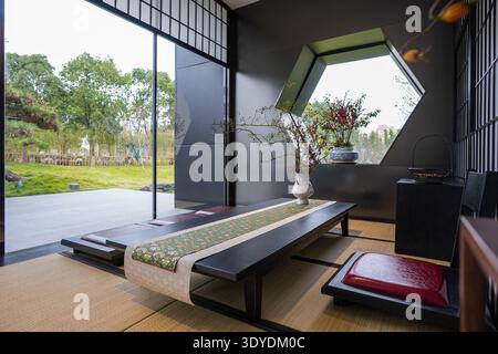 Vista dal pavimento tatami di una sala da tè Zen che guarda oltre un basso tavolo verso un lussureggiante giardino verde attraverso grandi finestre aperte. L'immagine trasmette il marinaio Foto Stock