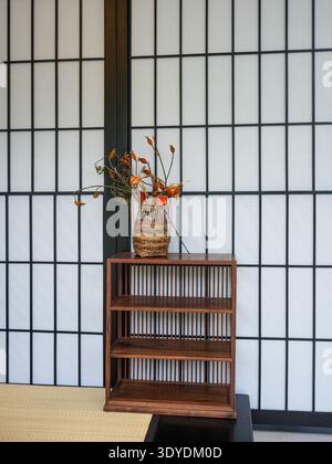 Piccolo scaffale in legno scuro che regge un vaso con rami secchi davanti a un tradizionale schermo shoji su un pavimento tatami. L'immagine trasmette una quantità minima Foto Stock