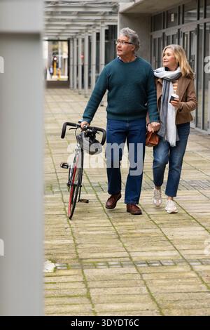 Coppia senior che cammina su un passaggio coperto, uomo che tiene la bicicletta, donna che porta una tazza di caffè e una borsa Foto Stock