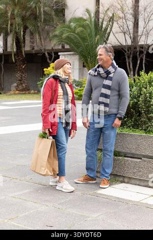 La coppia senior sta in piedi e sta conversando sul vialetto urbano pavimentato tenendo in mano borsa e zaino della spesa Foto Stock