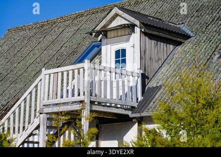 Un balcone in legno rustico con ringhiera bianca e porta si erge su un tetto intempestivo con muschio e licheni sotto un cielo azzurro limpido in una giornata di sole. Foto Stock