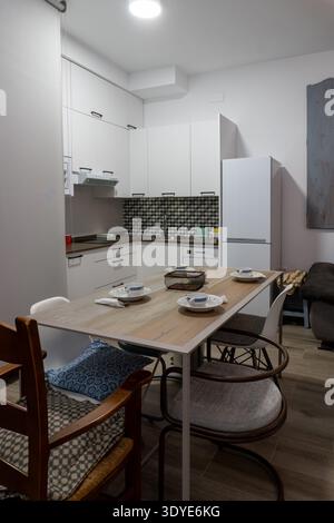 Cucina open space e sala da pranzo con tavolo in legno preparato per un pasto e frizzanti mobili bianchi Foto Stock