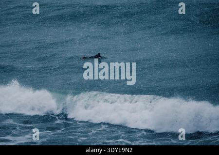Surfista pagaiando nell'oceano freddo durante le nevicate, onde potenti e sessioni di surf invernali. Foto Stock