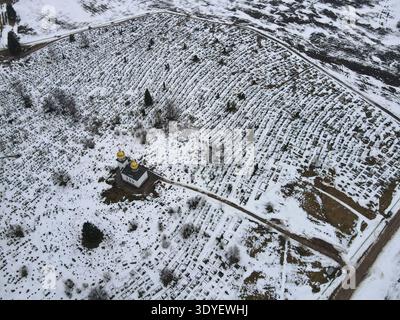 Vista aerea di un vasto cimitero bielorusso con file curvilinee di tombe innevate. Una piccola chiesa ortodossa con due cupole a cipolla dorate e tetto nero st Foto Stock