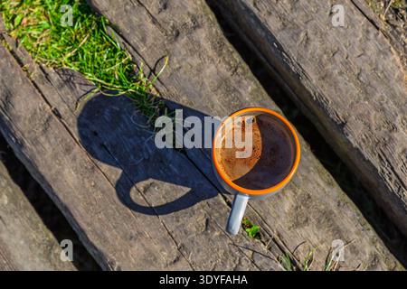 Una tazza di caffè in ceramica con bordo arancione in piedi su tavole di legno intemperie all'aperto. La schiuma è visibile sulla superficie della bevanda Foto Stock