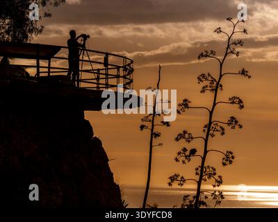 Il fotografo sagomato si erge su un punto panoramico della scogliera con un treppiede, catturando un tramonto di un'ora d'oro sull'oceano come spettacolari nuvole e piante costiere Foto Stock