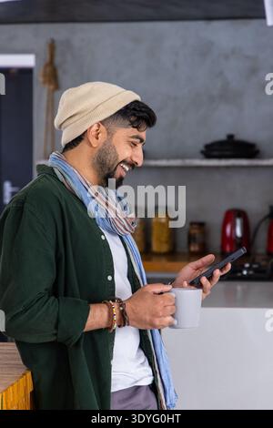 Uomo indiano che indossa berretto e sciarpa, sorride allo smartphone mentre tiene la tazza in ceramica in cucina Foto Stock