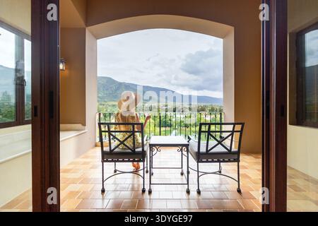 Una donna che indossa un cappello di paglia siede sul balcone di un hotel che guarda al paesaggio montano. Viaggi di lusso e relax in un resort panoramico con vista lago e medi Foto Stock