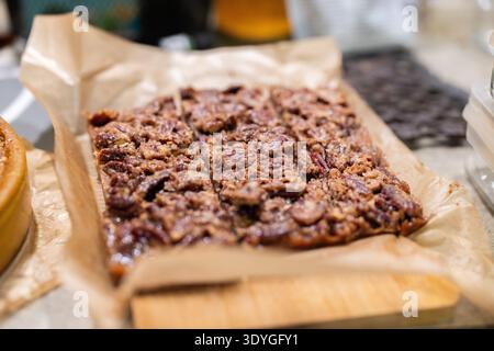 Le calde e gustose barrette di noci pecan poggiano su carta pergamena. Le fette mostrano noci caramellate ricche e bordi nitidi. Il dolce si trova invitante su una superficie di legno. P Foto Stock