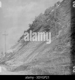 Fotografia vintage in bianco e nero che mostra una collina ripida e aspra con terreno esposto e alcuni alberi autunnali aggrappati al pendio. Pali di servizio in legno con linee elettriche corrono lungo la base della collina accanto a una strada sterrata, che illustra le infrastrutture rurali della metà del XX secolo. Il cielo coperto crea un paesaggio suggestivo e suggestivo, catturando l'ambiente naturale e il paesaggio quotidiano della regione. Uno sguardo nostalgico nel passato, che riflette l'atmosfera di un tranquillo Donbass prima della guerra. Foto Stock