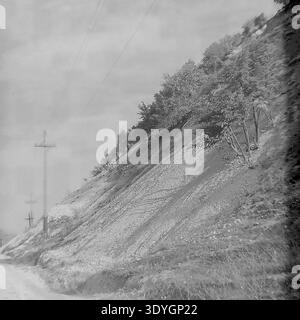 Fotografia vintage in bianco e nero che mostra una collina ripida e aspra con terreno esposto e alcuni alberi autunnali aggrappati al pendio. Pali di servizio in legno con linee elettriche corrono lungo la base della collina accanto a una strada sterrata, che illustra le infrastrutture rurali della metà del XX secolo. Il cielo coperto crea un paesaggio suggestivo e suggestivo, catturando l'ambiente naturale e il paesaggio quotidiano della regione. Uno sguardo nostalgico nel passato, che riflette l'atmosfera di un tranquillo Donbass prima della guerra. Foto Stock