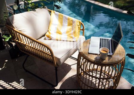 L'area salotto esterna presenta una sedia in rattan con una coperta a strisce, un computer portatile su un tavolo rotondo e una tazza da caffè accanto ad una piscina con vegetazione Foto Stock