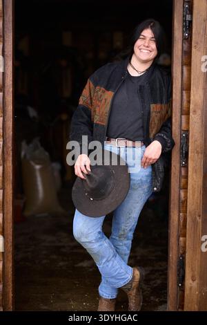 Una donna sorridente in un abbigliamento casual western si erge su una porta di legno, reggendo un cappello a teglia larga. Indossa jeans, una giacca a motivi geometrici e stivali, trasmettendo uno stile di vita rilassato e amichevole in un ranch. || modello rilasciato Foto Stock