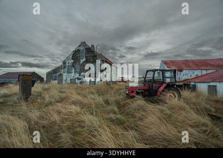 Edifici agricoli abbandonati e intemprati e vecchi trattori a Mýrar, Islanda occidentale, sotto un drammatico cielo coperto Foto Stock
