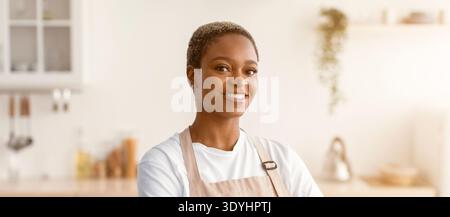 Una donna sorridente in un grembiule si trova in una cucina luminosa con un'atmosfera calda Foto Stock