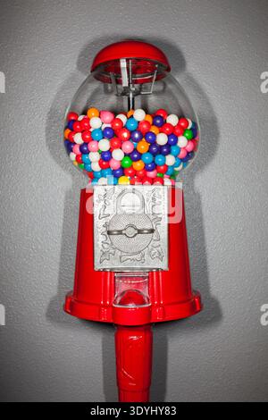 Primo piano di una macchina da pallone da masticare rossa piena di vibranti gomme multicolori contro una parete grigia. Concetto di caramelle divertenti e nostalgiche. Foto Stock