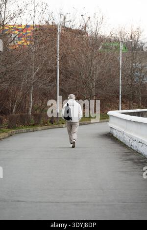 Vista posteriore di un giovane in una felpa con cappuccio bianca e cuffie che cammina da solo in un parco cittadino in una giornata nuvolosa. Stile di vita urbano moderno: Un uomo che indossa un filo Foto Stock