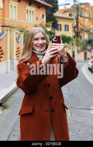 Donna sorridente che scatta foto con smartphone in un'affascinante strada europea con case colorate. Concetto di viaggio e stile di vita. Foto Stock