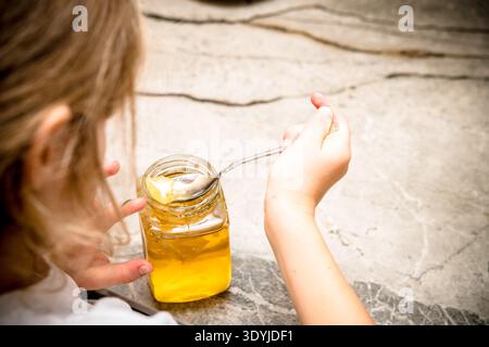 Primo piano delle mani di un bambino, tenendo un cucchiaio e raccogliendo il miele da un vaso di vetro su una superficie di pietra, preparando il tiglio alle erbe. Foto Stock