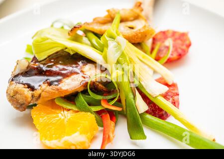 Un primo piano di carne alla griglia condita con salsa, servita con fette d'arancia, fette d'arancia e varie verdure verdi su un piatto bianco. Foto Stock