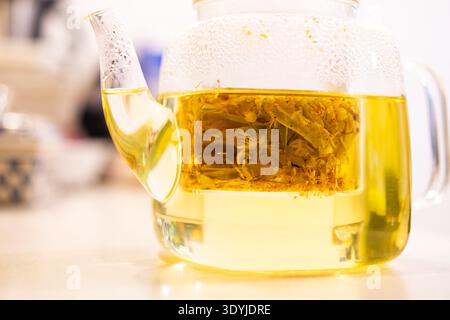 Teiera in vetro con acqua limpida che prepara fiori di tiglio naturali per tè alle erbe su una superficie leggera con sfondo sfocato. Foto Stock