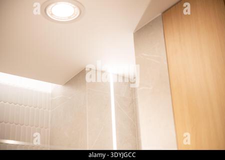 Vista ravvicinata di un moderno angolo bagno con plafoniera incassata, piastrelle in marmo beige, striscia luminosa LED verticale e pannelli in legno. Foto Stock