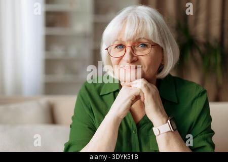 Donna anziana con gli occhiali che appoggiano il mento sulle mani mentre è seduta a casa Foto Stock