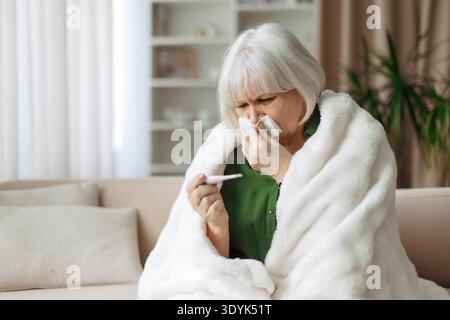 Donna anziana che controlla la temperatura corporea con un termometro mentre è avvolta in coperta Foto Stock