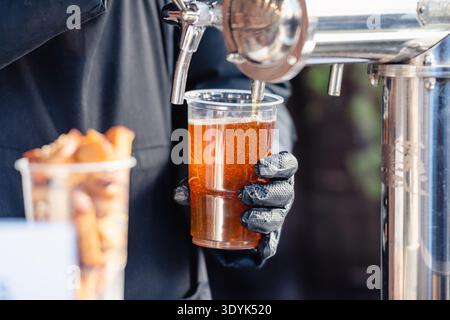 Primo piano di un barista che versa birra alla spina fresca in una tazza di plastica durante un festival all'aperto. Una lager fresca e schiumosa viene servita da una spina Foto Stock