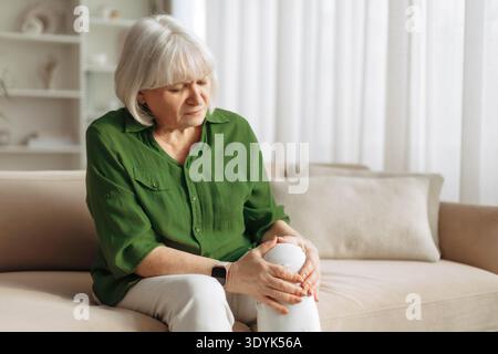 Donna anziana che tiene il ginocchio doloroso seduto sul divano a casa Foto Stock